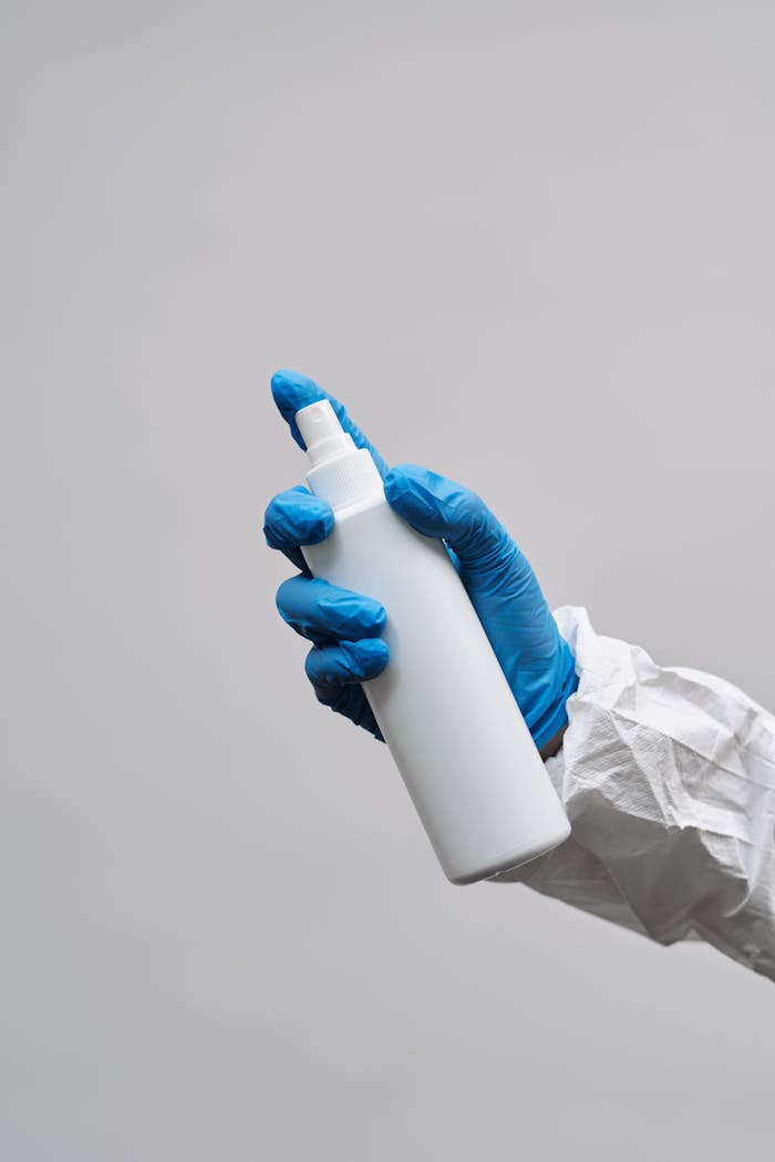 Close-up of a blue-gloved hand holding a white spray bottle against a light gray background.